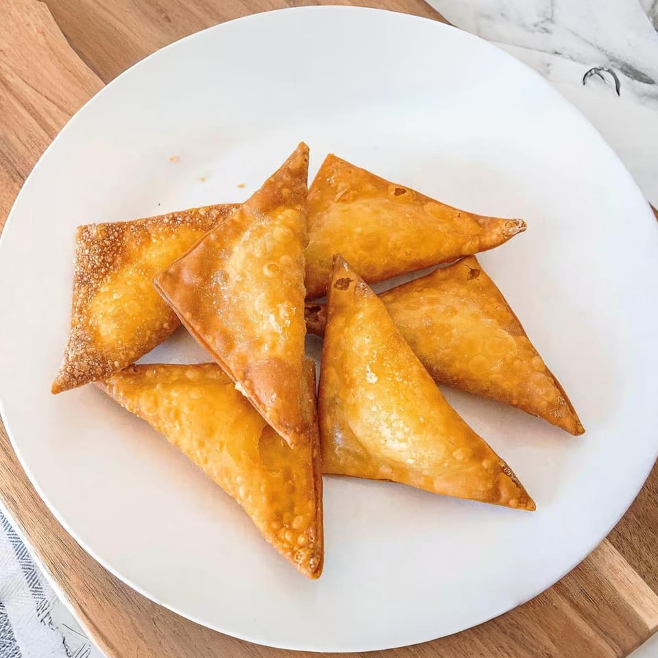 A white plate holds six golden-brown, triangular fried wontons, ready to be served.